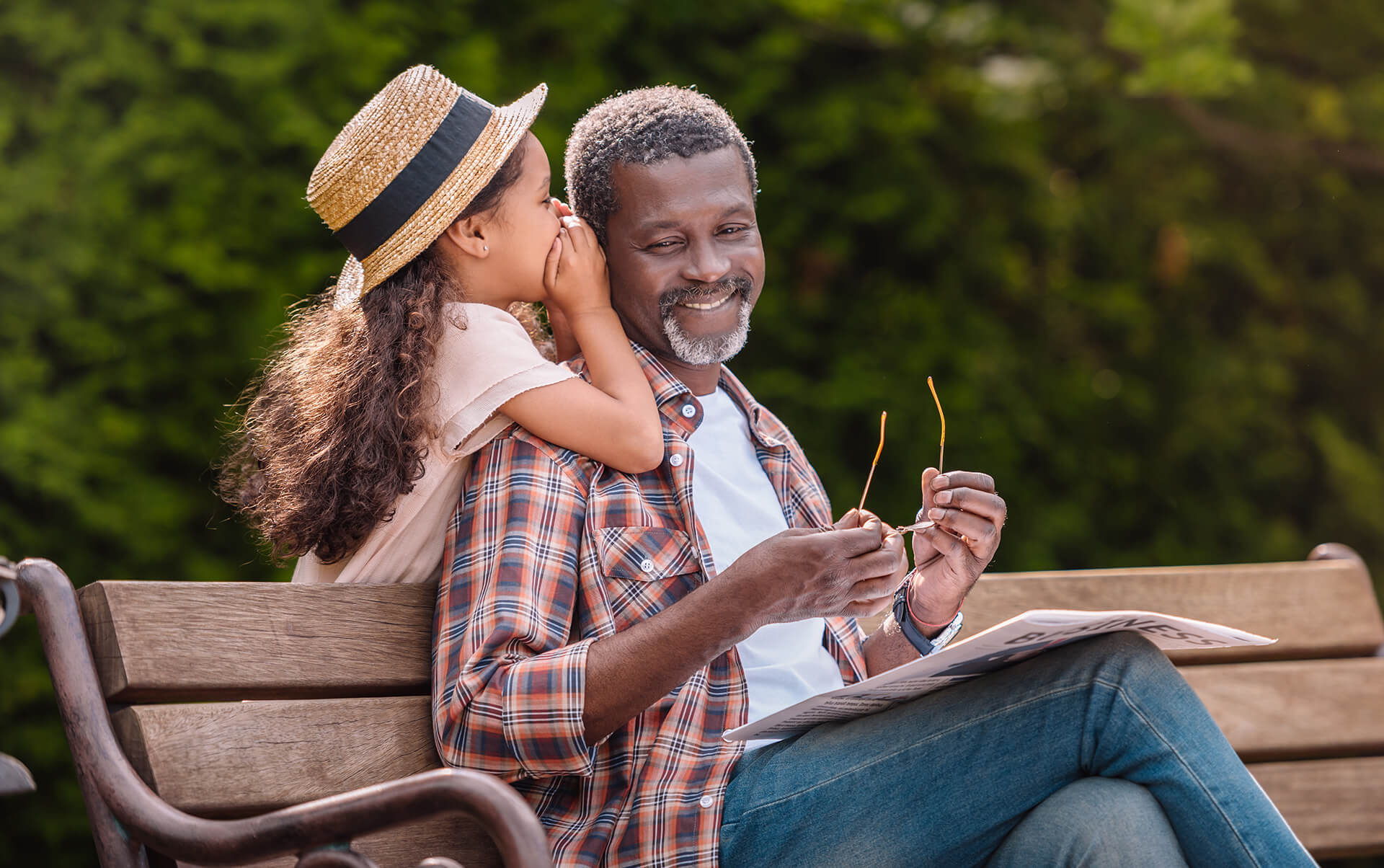 A grandfather relaxing on a park bench while his granddaughter whispers in his ear