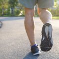 Close-up of a person walking or jogging on a paved path, wearing athletic shoes and shorts, in a sunny park setting.