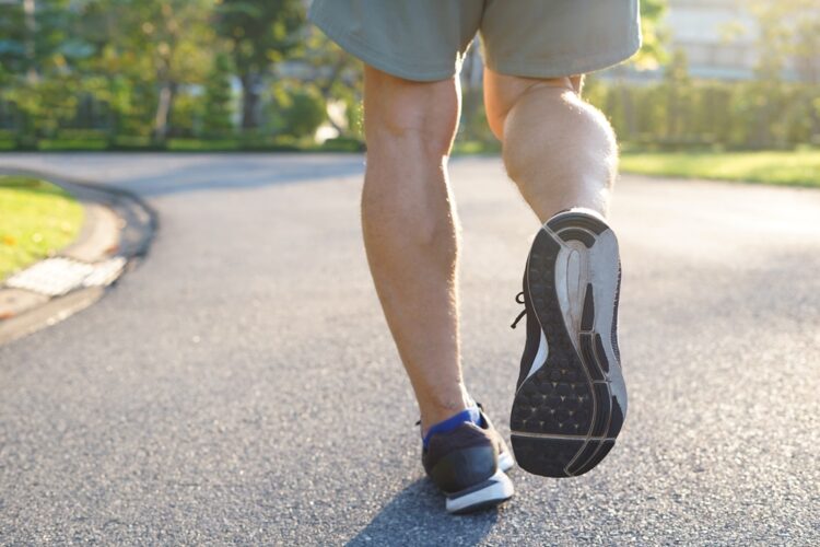 Close-up of a person walking or jogging on a paved path, wearing athletic shoes and shorts, in a sunny park setting.