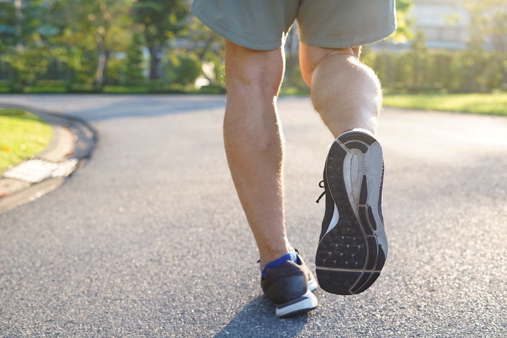 Primer plano de una persona caminando o trotando en un camino pavimentado, vistiendo zapatos deportivos y pantalones cortos, en un parque soleado.